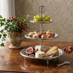 Könnte beinhalten: Ein dreistöckiges Serviertablett aus Silber mit Trauben, Gebäck, Käse, Feigen und Nüssen. Eine Topfpflanze mit grünen Blättern und roten Beeren steht in der Nähe auf einem Holztisch. Ein dekorativer Schlüssel liegt ebenfalls auf dem Tisch.