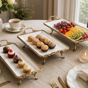 May include: Rectangular serving platters with gold-coloured handles and legs. The trays display pastries and fresh fruit, such as grapes, strawberries, oranges, and kiwi. A lit candle and a cup of tea are also present.