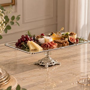 Peut inclure: Un plateau de service rectangulaire en argent avec un bord perlé et une base sur pied décorative, contenant un assortiment de fromages, raisins, figues et craquelins. Le plateau présente une surface réfléchissante et un pied en métal orné.