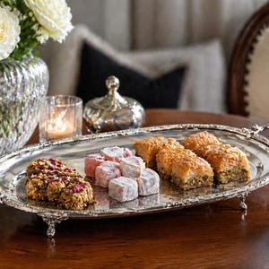 Op de afbeelding: Een ovale zilveren serveerschaal met sierlijke handvatten en voetjes presenteert een assortiment gebak, waaronder baklava en Turks fruit. Een kopje koffie staat in de buurt, met een vaas met witte rozen op de achtergrond.