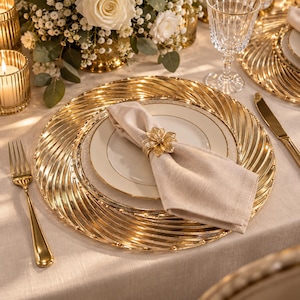 Pode incluir: Um prato de serviço dourado com um padrão radial em espiral, coberto por um prato branco com uma borda dourada fina. Um guardanapo de linho bege está dobrado e preso com um anel de guardanapo dourado metálico em forma de flor, ao centro.