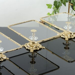 May include: A set of three rectangular glass display stands with gold-toned metal frames and ornate, decorative gold bases. Each stand features a clear glass top supported by a central pedestal with a crystal-like accent above the base.