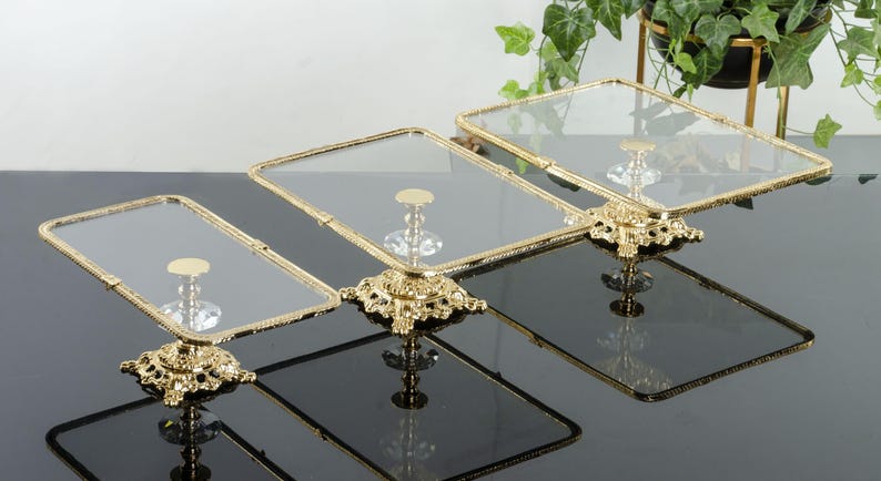 May include: A set of three rectangular glass display stands with gold-toned metal frames and ornate, decorative gold bases. Each stand features a clear glass top supported by a central pedestal with a crystal-like accent above the base.