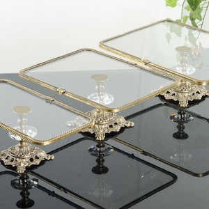 May include: A set of three rectangular glass display trays with brass-toned metal frames. Each tray features a clear glass surface and is supported by an ornate, pedestal-style base with intricate metal detailing and a central clear glass accent.