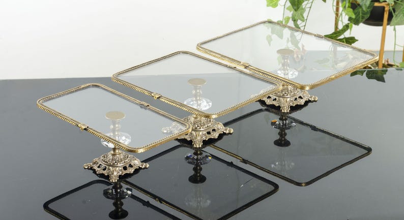 May include: A set of three rectangular glass display trays with brass-toned metal frames. Each tray features a clear glass surface and is supported by an ornate, pedestal-style base with intricate metal detailing and a central clear glass accent.