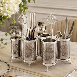 May include: A silver-toned metal cutlery caddy featuring five cylindrical containers with ornate floral engravings. The caddy includes two vertical handles with circular loops at the top and stands on small, decorative feet, holding various forks, spoons, and knives.