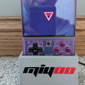 May include: A handheld gaming console with a purple translucent case, black directional pad, and pink buttons. The screen displays a red triangle on a purple background. The console sits in a gray stand with the text "qiyoo" in black and red.