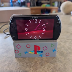 May include: A black Sony PSP device displaying a clock face, resting on a gray stand. The stand features colorful PlayStation symbols and a stylized "P" logo. The clock shows the date "MON 13".