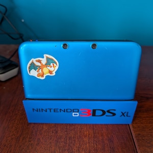 May include: Blue Nintendo 3DS XL handheld game console with a Charizard sticker. The console is on a blue stand with the text "NINTENDO 3DS XL". The console has two camera lenses.