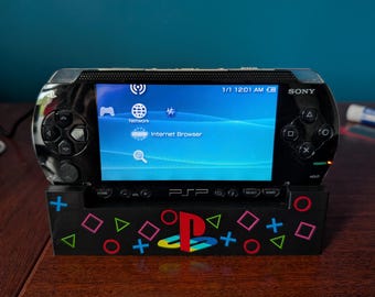 May include: A black Sony PSP handheld gaming console resting in a custom black stand. The screen displays the main menu with Network and Internet Browser icons. The stand features colorful PlayStation controller symbols including blue plus signs, pink squares, green triangles, and red circles, with a central PlayStation logo.