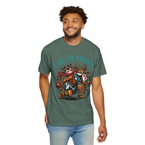 May include: Green t-shirt featuring a cartoon illustration of a figure with a deer head, holding maracas. The figure is wearing a red scarf and a white skirt. The text "LIOS EM CHANIA" is arched above the figure in teal.