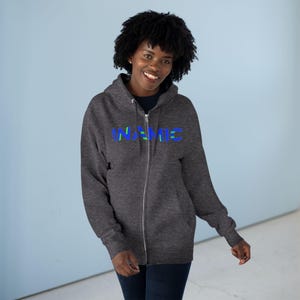 Könnte beinhalten: Ein dunkel anthrazitfarbener Kapuzenpullover mit Reißverschluss, Kapuze mit Kordelzug und langen Ärmeln. Auf der Vorderseite ist das Wort INAMIC in einer kräftigen, stilisierten Schrift mit blauen und grünen horizontalen Akzenten gedruckt.