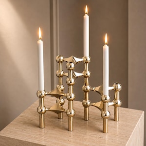 Puede incluir: Un candelabro de metal dorado con un diseño modular de acentos esféricos apilados y conectores puntiagudos. La estructura sostiene tres velas blancas encendidas en soportes verticales, creando una disposición escalonada de superficies.