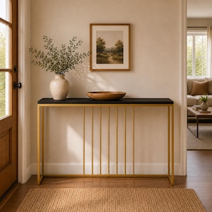 May include: A console table featuring a rectangular black top and a gold-toned metal frame with vertical bars. On the table sits a beige speckled vase filled with green leafy branches and a large, shallow wooden bowl centered on the surface.