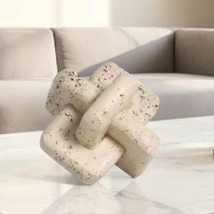 May include: Cream-colored decorative knot sculpture with dark speckles. The interlocking design features rounded rectangular shapes, creating a modern, abstract art piece. This home decor item adds a touch of elegance to any space.