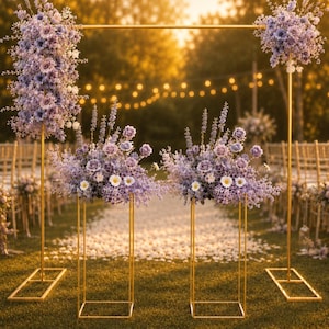 May include: A golden metal wedding arch and floral stands are adorned with lavender and white flowers. The arch is square, and the stands have a geometric design. The scene is set outdoors with a blurred background of trees and string lights.