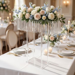 May include: A tall, rectangular clear acrylic flower stand featuring a prism-like border design. The stand is topped with a lush arrangement of white and pale pink roses mixed with light blue delphinium flowers and green foliage.