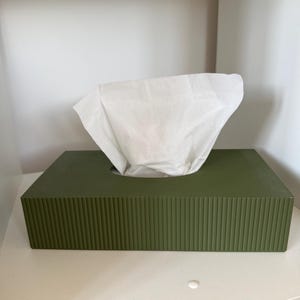 May include: A rectangular olive green tissue box cover featuring a vertical ribbed texture on all sides. A single white paper tissue is pulled through the oval opening at the top, extending upwards from the centre of the solid green container.