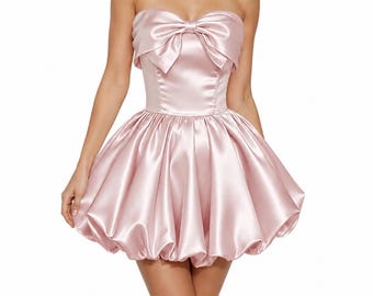 May include: A strapless, light pink satin dress. The bodice features a large bow at the bust, and the skirt is a voluminous bubble shape. The dress is designed to be form-fitting and is made of a shiny, smooth fabric.