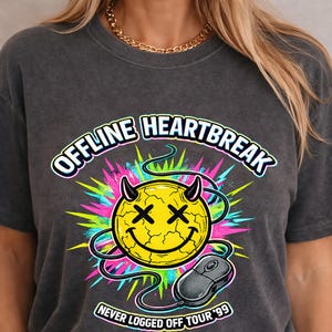 May include: Dark grey t-shirt with a graphic design featuring a yellow smiley face with devil horns and crossed-out eyes. The text "OFFLINE HEARTBREAK" and "NEVER LOGGED OFF TOUR '99" are also displayed.
