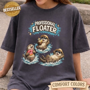 May include: Dark grey t-shirt with the text "PROFESSIONAL FLOATER" above an illustration of three otters floating in water. One otter holds a donut, another a coconut drink, and the third is relaxing. The shirt has a "BESTSELLER" and "COMFORT COLORS" label.