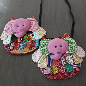 Op de afbeelding: Twee handgemaakte olifantvormige schoudertassen. Elke tas heeft een roze olifantenkop met stoffen oren en een patchwork-lichaam in verschillende kleuren en patronen. De tassen hebben een zwarte riem om te dragen.