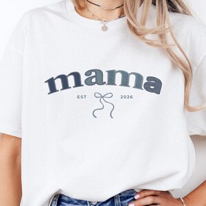 Könnte beinhalten: Weißes T-Shirt mit dem Wort "mama" in dunkelblauer Schrift in Bogenform. Darunter steht "EST 2026" und eine Schleifen-Grafik. Das Shirt wird mit blauen Jeansshorts kombiniert.