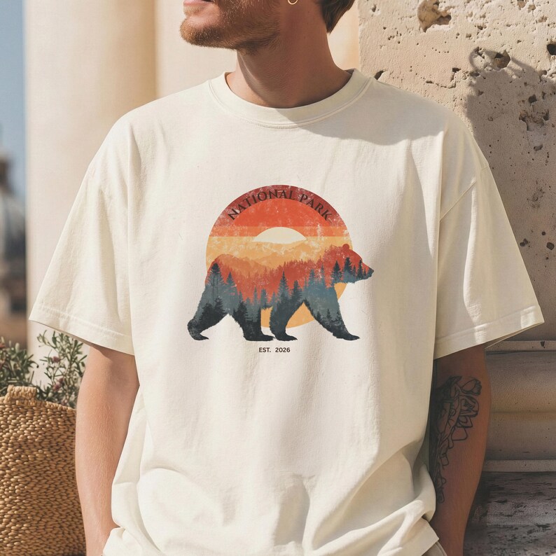 May include: Cream-colored t-shirt featuring a circular graphic with a bear silhouette and the words "NATIONAL PARK" in a semi-circle above the bear. The graphic includes a sunset design with trees and the text "EST. 2026".