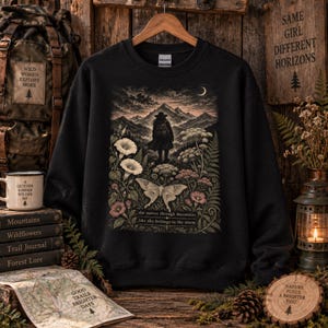 Peut inclure: Un sweat-shirt noir à col rond orné d'une illustration représentant une silhouette face à une chaîne de montagnes sous un croissant de lune. Le motif inclut des fleurs, des fougères, un grand papillon de nuit et le texte : she moves through mountains like she belongs to the storm.