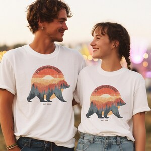May include: White t-shirts with a graphic of a bear in a forest scene, set against a sunset. The text "NATIONAL PARK" arches above the bear, with "EST. 2026" below. The shirts are worn by two people.