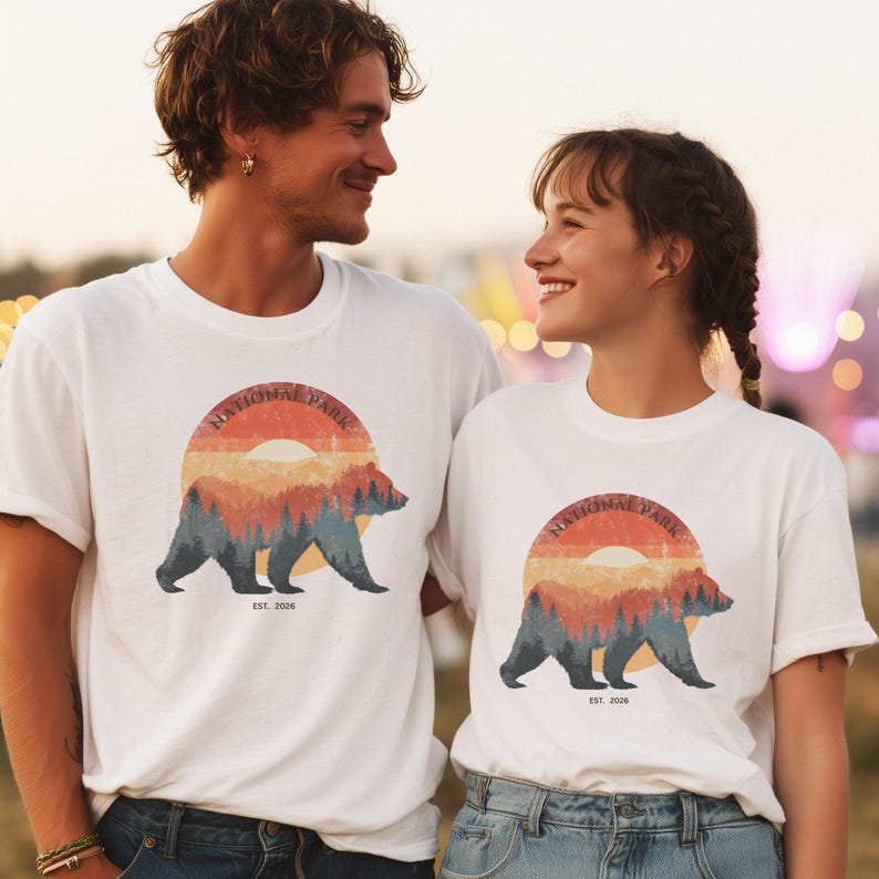 May include: White t-shirts with a graphic of a bear in a forest scene, set against a sunset. The text "NATIONAL PARK" arches above the bear, with "EST. 2026" below. The shirts are worn by two people.