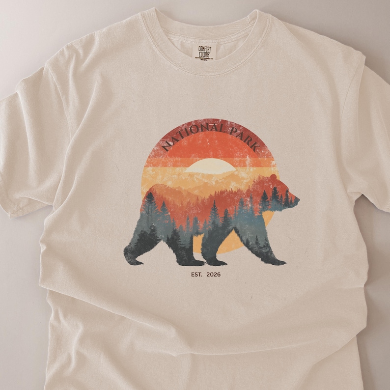 May include: Cream-colored t-shirt featuring a bear silhouette with a forest scene inside a circular design. The design includes the words "NATIONAL PARK" and "EST. 2026". The bear is in front of a sunset-colored background.
