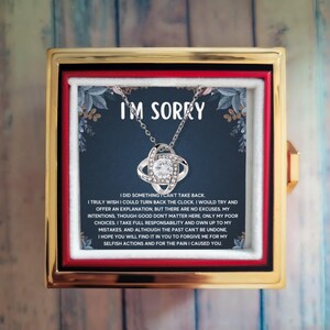 May include: A silver-toned necklace with a central knot pendant featuring a clear round stone, presented in a gold-colored box. The card inside displays the text I'M SORRY followed by a paragraph expressing regret and a request for forgiveness.