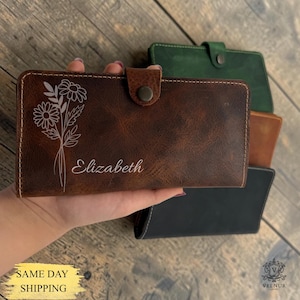 May include: A brown leather wallet with a floral design and the name "Elizabeth" engraved on the front. The wallet has a button closure and is surrounded by other wallets in green, tan, and black.