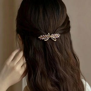 May include: A gold-coloured hair clip with a leaf and bow design, embellished with purple-pink gemstones. The clip is set against dark brown hair, with the back of a person's head visible. The hair is styled in loose waves, with the clip securing a section of the hair.
