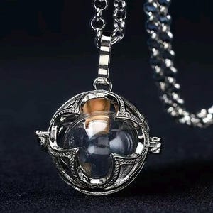 May include: A silver-colored pendant necklace featuring a clear glass orb with a cork stopper, encased in an ornate, openwork metal cage. The pendant hangs from a delicate chain. The background is a dark, solid color.