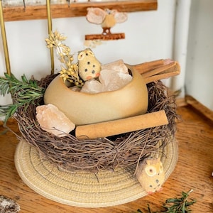 Pode incluir: Arranjo decorativo com tema de coruja. Uma tigela de cerâmica cheia de cristais e paus de canela em um ninho tecido. Inclui uma pequena estatueta de coruja e galhos verdes, tudo em uma base tecida. Design rústico.