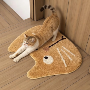 Puede incluir: Una alfombra en forma de gato de color beige con un diseño de cara de oso de dibujos animados. La alfombra está hecha de un material suave y afelpado. Un gato atigrado se estira sobre la alfombra.