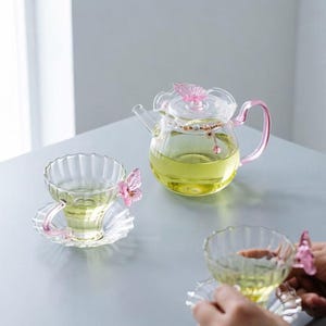 May include: A clear glass teapot with a pink butterfly on the lid and a pink handle, filled with light yellow tea. Two matching fluted glass teacups with pink butterfly accents on the handles sit on matching saucers, also containing yellow tea.