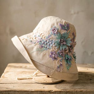 May include: A beige bucket hat featuring intricate floral embroidery in shades of lavender, mint green, and soft blue. The design includes three-dimensional fabric flower appliqués and small white pearl-like beads scattered across the side.
