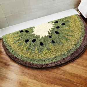 May include: A half-circle rug designed to resemble a kiwi fruit. The rug features shades of green, brown, and white, with black seed details. The rug is placed on a wooden floor.