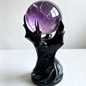 May include: A translucent purple sphere with a network of white internal fractures held by a black, glossy, hand-shaped stand. The stand features elongated, pointed fingers that cradle the orb, with a smooth, swirling base design.