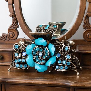 May include: A decorative hair clip featuring a large turquoise flower design with a central cluster of smaller blue gems. The clip has a bronze-coloured metal frame with leaf and gem accents. The clip is resting on a wooden surface.