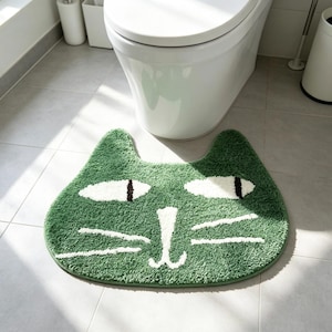 Peut inclure: Un tapis de bain vert en forme de tête de chat avec des yeux blancs et noirs, un nez blanc et des moustaches blanches. Le tapis présente une surface texturée et touffetée, avec deux oreilles pointues sur le dessus, formant une silhouette féline.