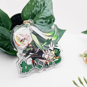 Puede incluir: Un charm de acrílico transparente con un personaje de dibujos animados con cabello blanco y detalles verdes, sosteniendo un puerro y acompañado por una criatura parecida a un pájaro. El charm tiene un llavero dorado y texto japonés.