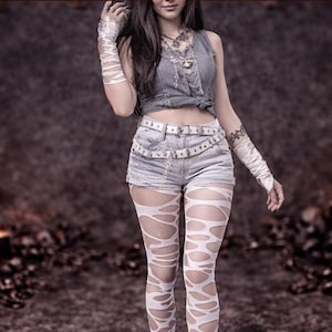 Puede incluir: Un top corto de mezclilla gris y pantalones cortos de mezclilla con un cinturón blanco. El atuendo se combina con medias blancas rasgadas y envolturas para los brazos. El fondo es una superficie oscura y texturizada con velas y calaveras.