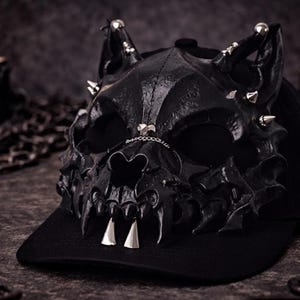 May include: Black baseball cap with a sculpted skull mask. The mask has sharp teeth, silver spikes, and a chain detail. The cap is set against a dark background with candles and chains, creating a gothic aesthetic.