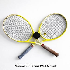 May include: Two yellow tennis rackets with black strings are crossed, with a black ball in the center. The handles are wrapped in gray and brown grips. A yellow tennis ball is visible. Text at the bottom reads "Minimalist Tennis Wall Mount."