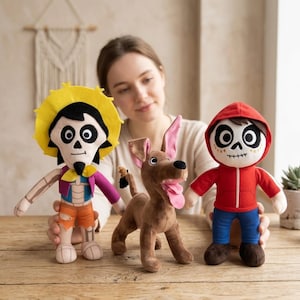 Può includere: Tre bambole di peluche ispirate al film d'animazione Coco. Le bambole raffigurano un personaggio con un cappello giallo, un cane marrone e un personaggio con una felpa rossa. Le bambole sono su una superficie di legno.