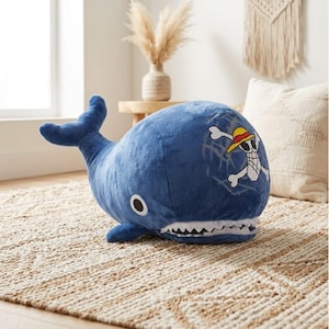 May include: A plush blue whale-shaped pillow with a white mouth and teeth. The pillow features a cartoon skull and crossbones design on its side. The whale is resting on a beige and white patterned rug.